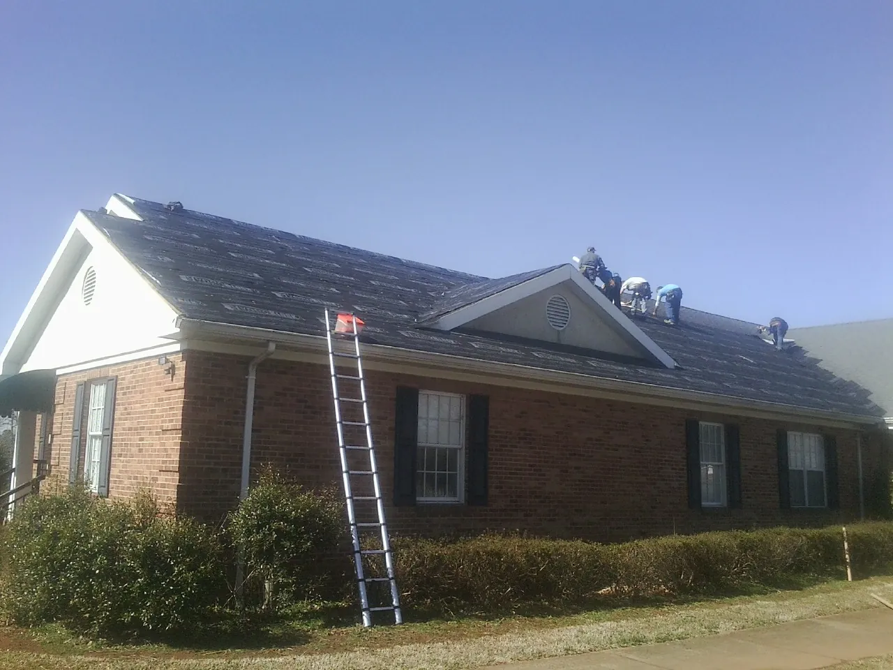 Crew tearing shingles off a brick ranch roof with a ladder set — Emergency Roof Repair work by our crew in Combined Locks